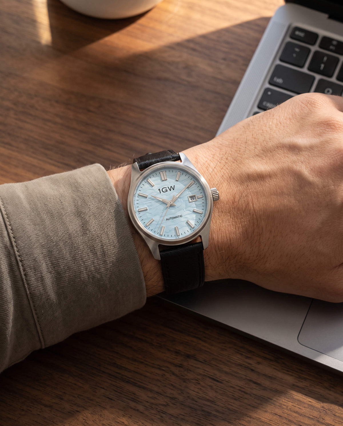 Ice-blue textured dial automatic watch with steel case and black leather strap, worn on a wrist at a desk beside a laptop